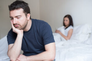 Man sitting on the edge of a bed looking stressed over possible erectile dysfunction with woman sitting in background